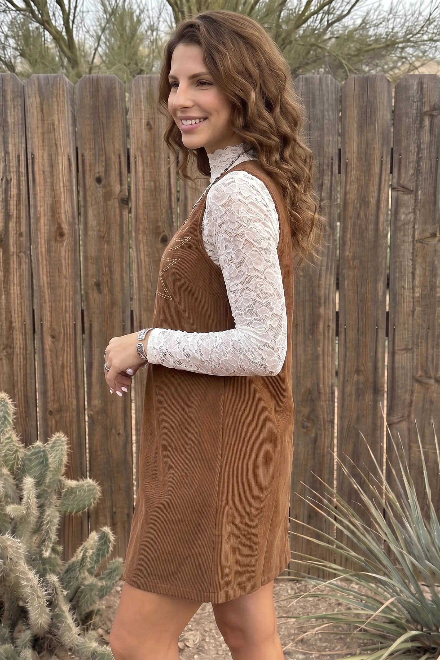 A woman wearing a brown corduroy pinafore dress over a white lace long-sleeve top, standing outdoors in front of a wooden fence with desert plants.