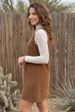 A woman wearing a brown corduroy pinafore dress over a white lace long-sleeve top, standing outdoors in front of a wooden fence with desert plants.