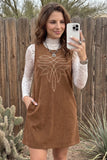 A woman takes a mirror selfie wearing a brown corduroy sleeveless dress over a white lace long-sleeve turtleneck. The dress features a white embroidered geometric design on the chest and side pockets.