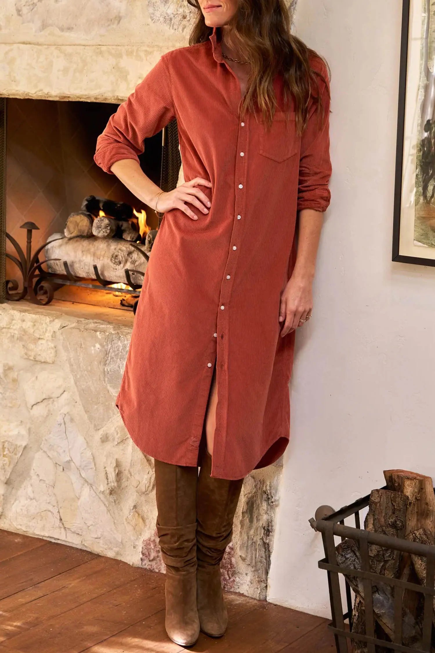 A woman models a rust-colored long button-up shirt dress with a chest pocket, paired with brown suede boots, standing in front of a stone fireplace.
