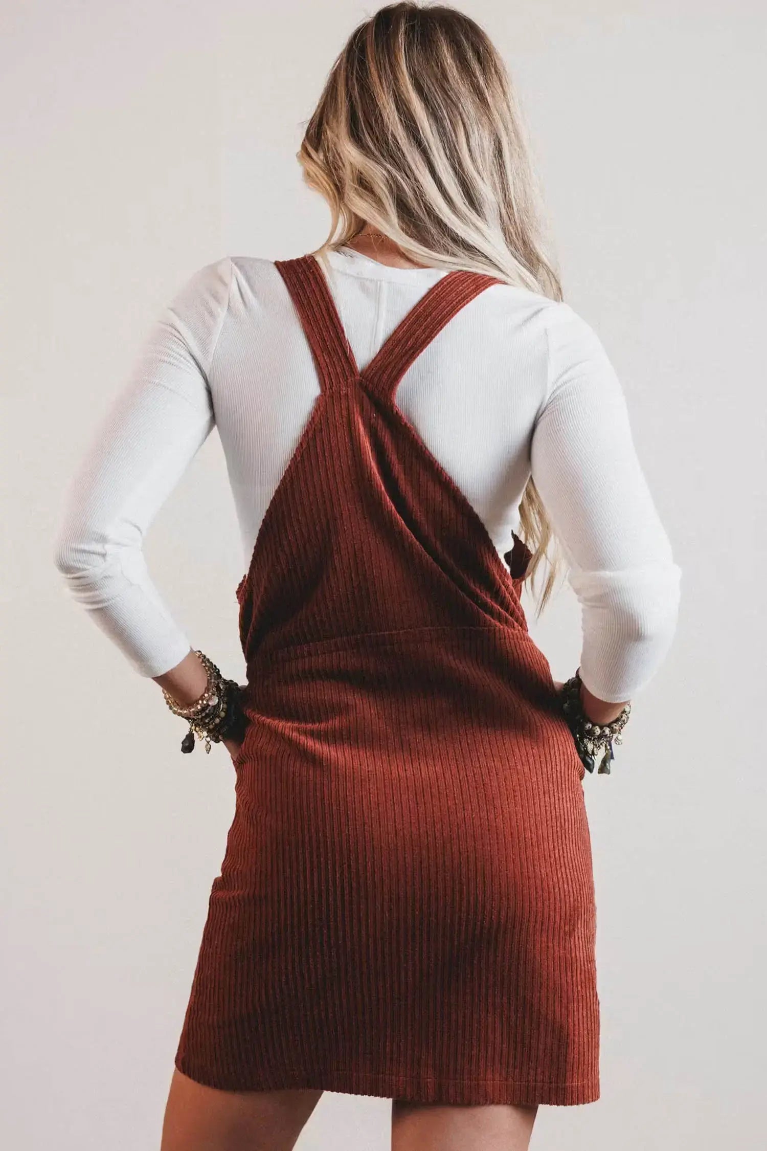 A woman shown from the back wearing a rust-brown ribbed corduroy pinafore dress over a white long-sleeve shirt, set against a plain light gray background.