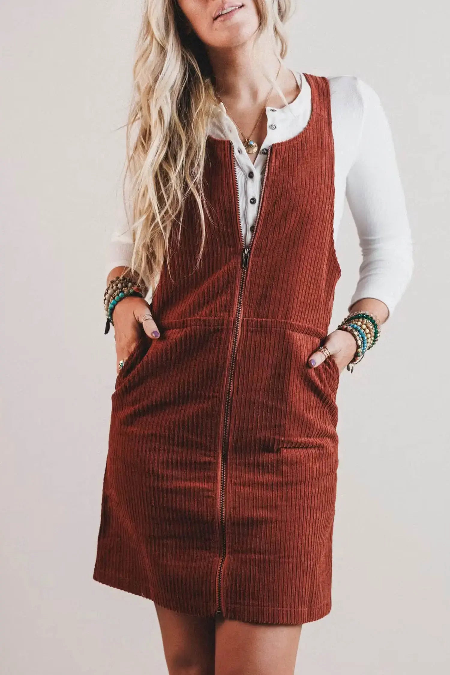 A woman wearing a rust-brown corduroy zip-front pinafore dress over a white collared shirt, standing against a plain light beige background.