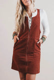 A woman wearing a rust-brown corduroy zip-front pinafore dress over a white collared shirt, standing against a plain light beige background.