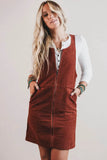 A woman wearing a rust-brown corduroy pinafore dress over a white long-sleeve button-up shirt, standing against a plain light gray background.