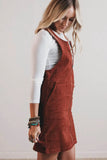A woman wearing a rust-brown corduroy pinafore dress over a white long-sleeve top, standing against a plain off-white wall.