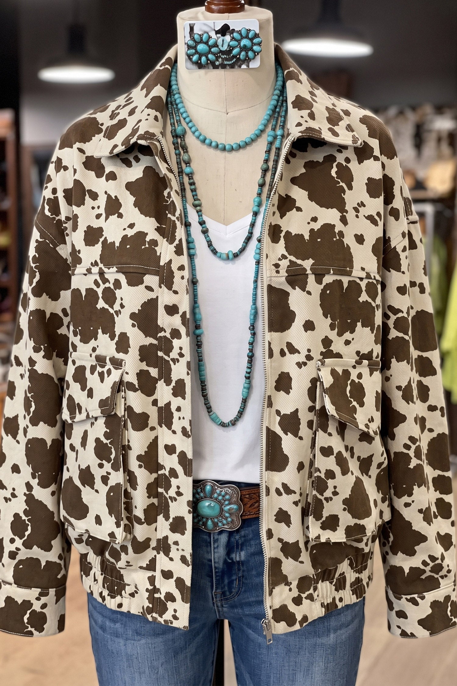 Cow-print jacket on mannequin styled with white top, blue jeans, turquoise necklaces, and belt buckle.