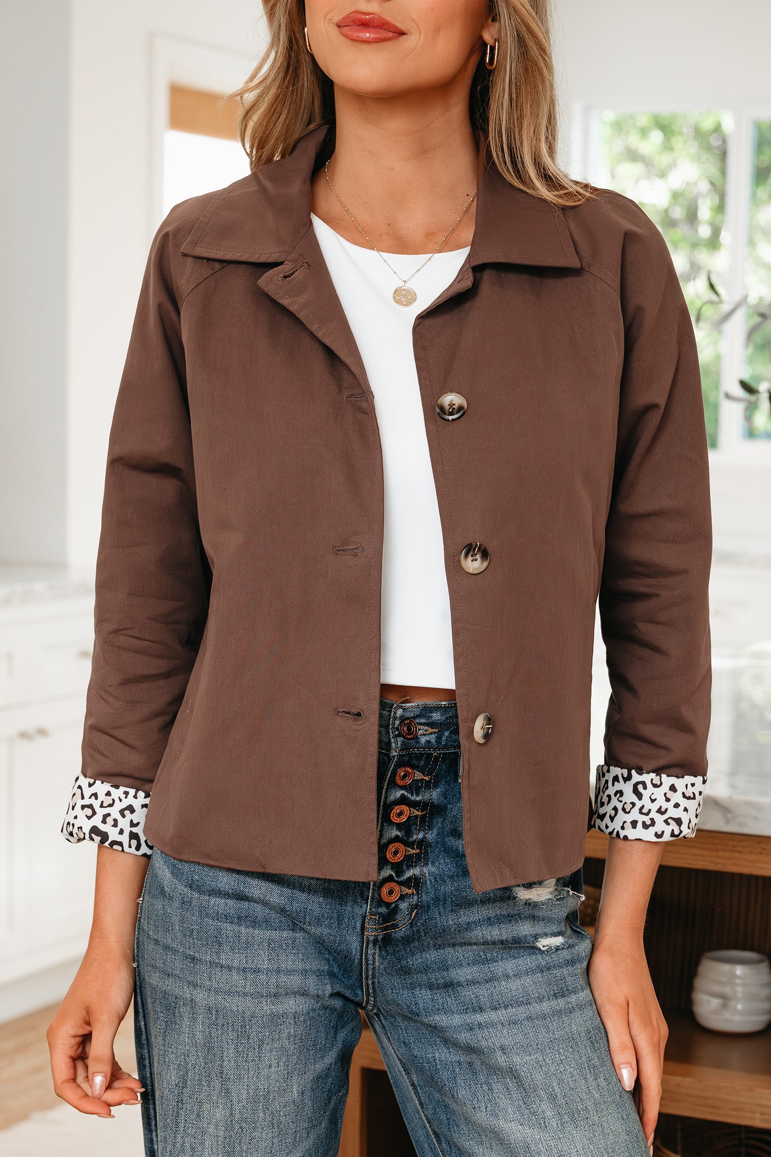 Woman wearing a brown jacket with button details, white shirt, and blue jeans indoors.