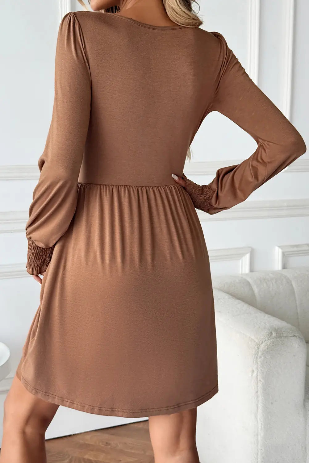 A woman wearing a long-sleeve brown dress, shown from the back in an indoor setting with a white sofa in the background.