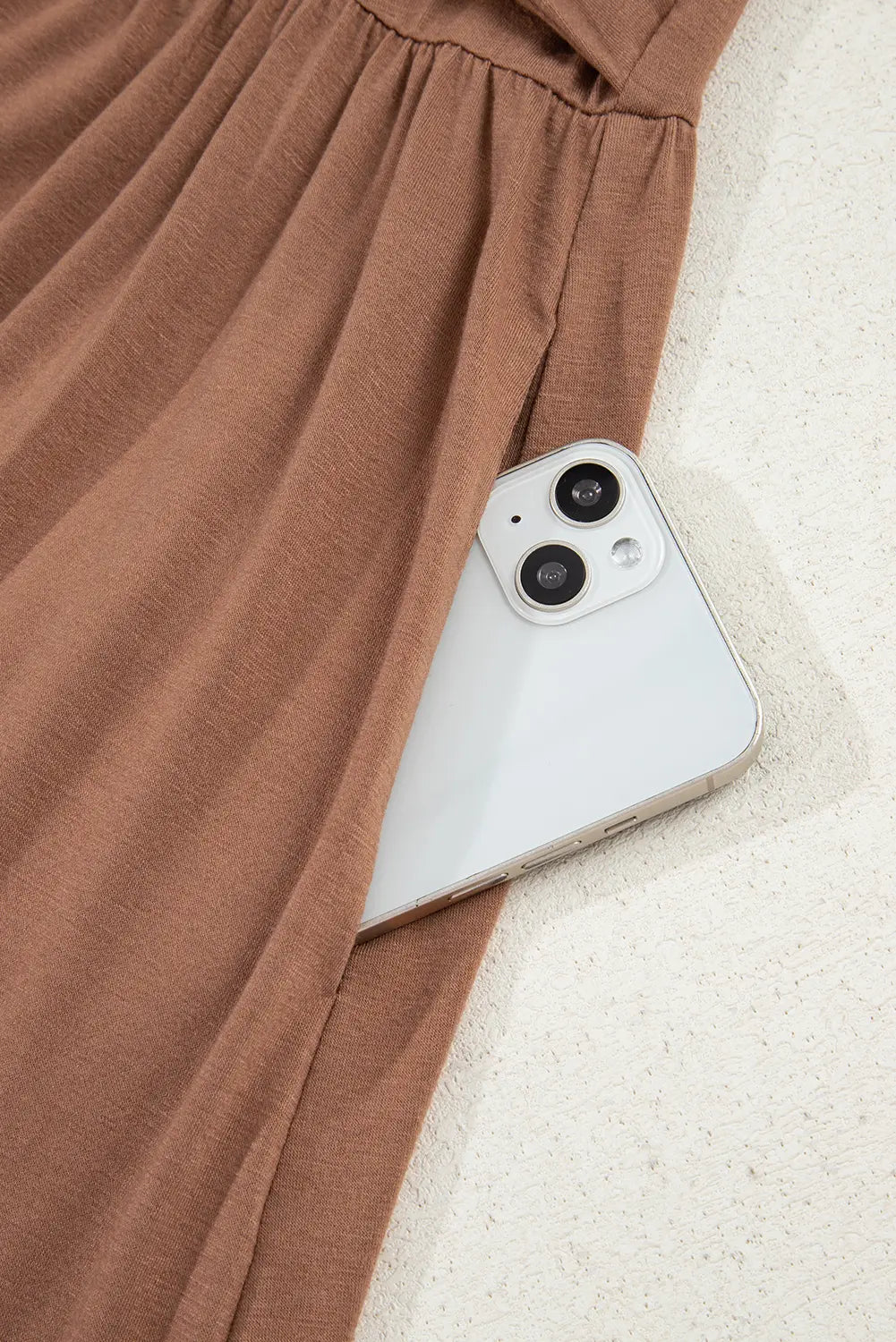A close-up of a brown draped garment with a side pocket, shown holding a white smartphone on a light textured surface.