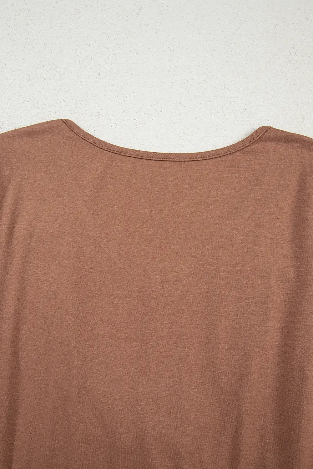A close-up of a brown crew-neck t-shirt hanging against a textured off-white wall.