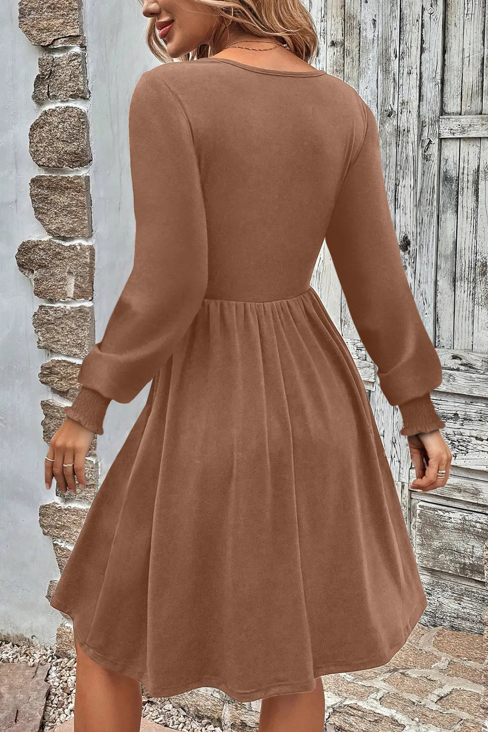 A woman wearing a long-sleeve brown dress, shown from the back in an outdoor setting with a rustic stone wall and weathered wooden door.