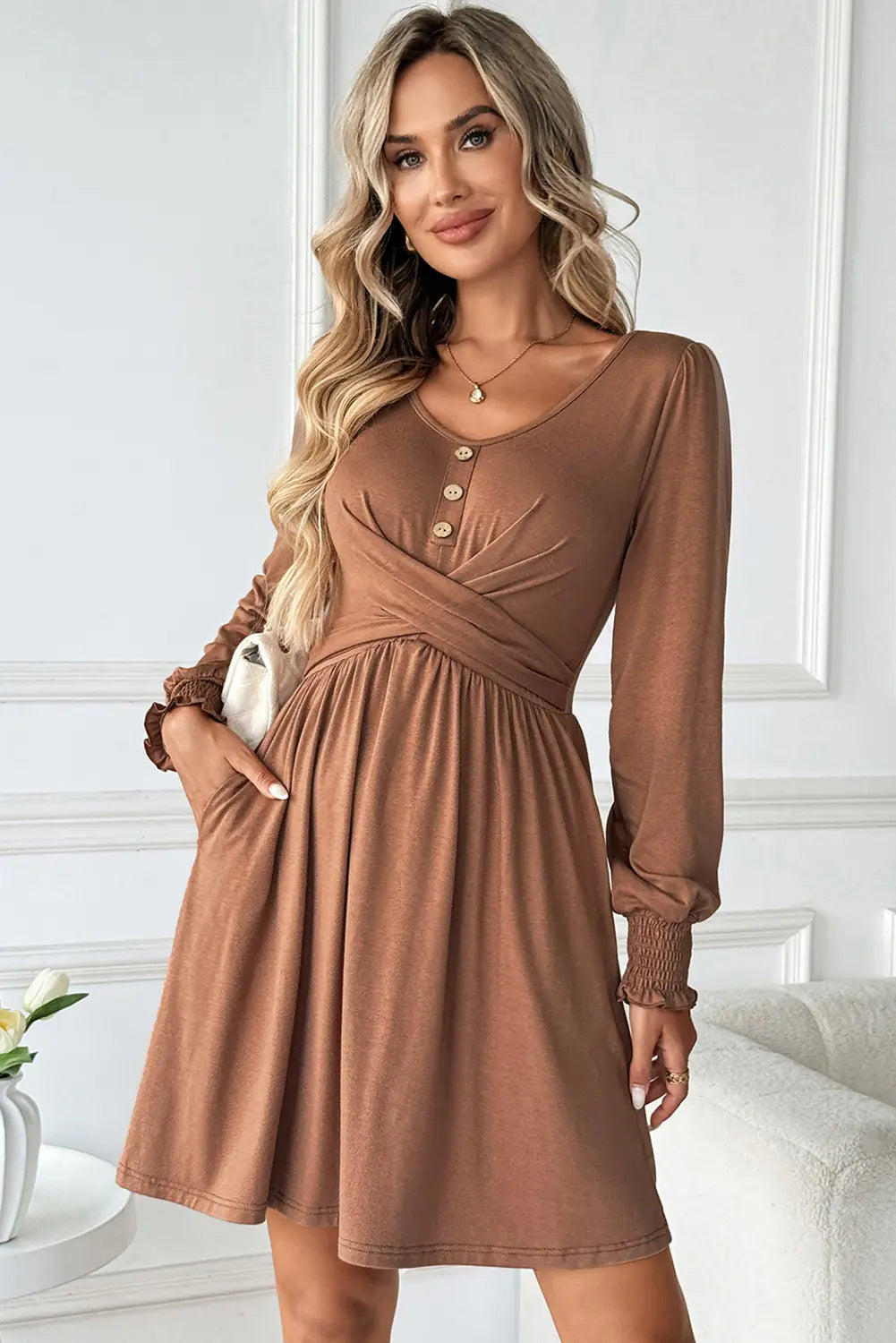A woman wearing a brown long-sleeve maternity dress with a crossover front, button details, and side pockets, posed in a bright indoor setting.