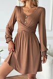 A woman wearing a brown long-sleeve mini dress with a V-neck, button detail at the chest, and a gathered waist, posed in an indoor setting.