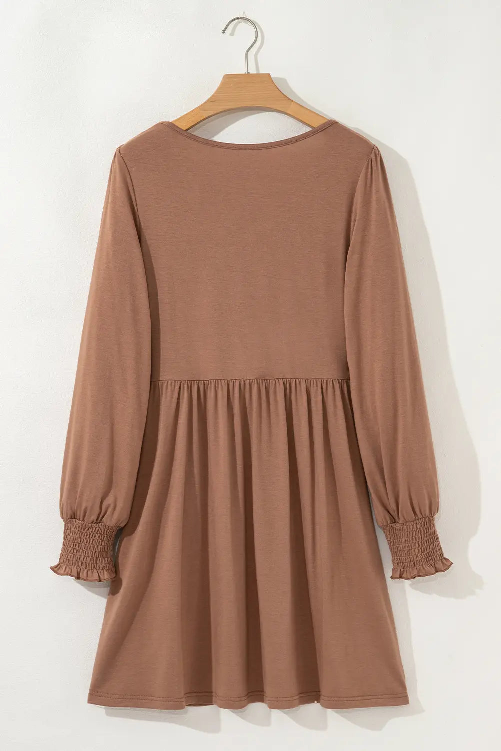 A long-sleeve brown dress with a gathered waist and smocked cuffs, hanging on a wooden hanger against a white wall.