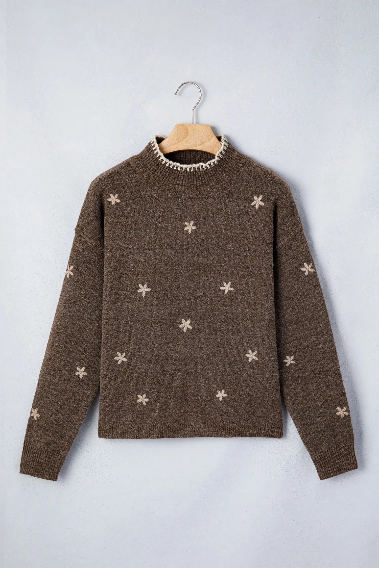 A brown knit sweater with small beige snowflake patterns, hanging on a wooden hanger against a light gray background.