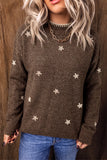 A woman wearing a brown knit sweater with small white floral motifs, paired with blue jeans and a gold chain necklace, standing against a dark wood background.