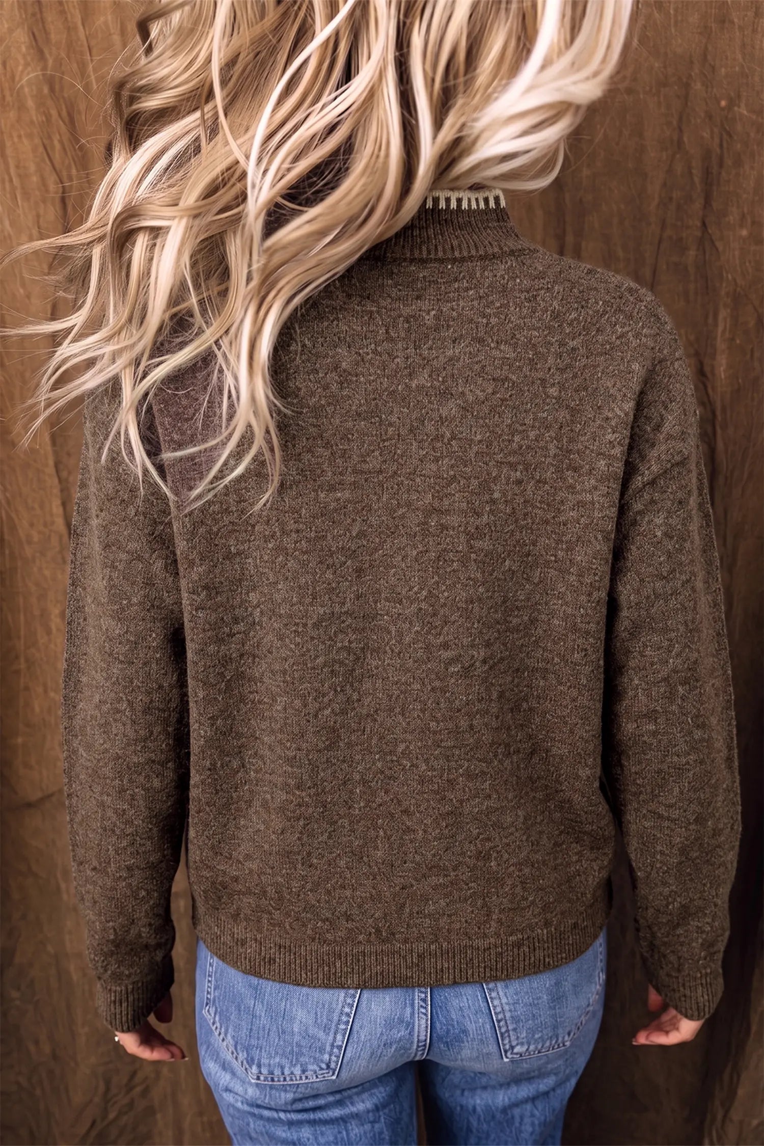A person viewed from the back wearing a brown knit sweater and blue jeans, standing against a brown textured backdrop.