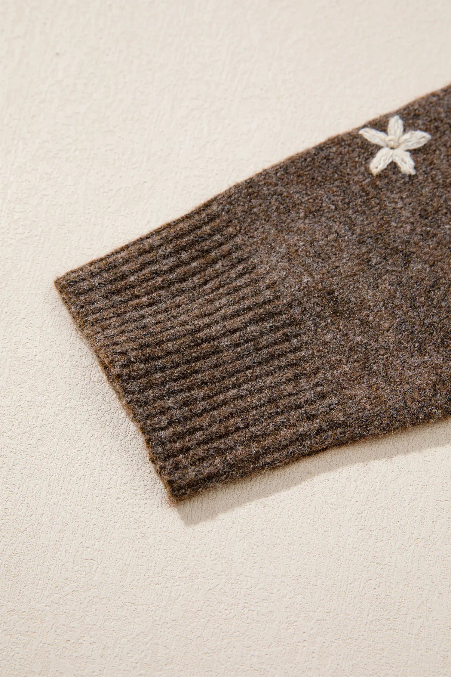 Close-up of a ribbed brown knit sleeve with a small white embroidered flower detail, set against a warm off-white textured background.