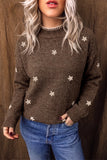 A woman wearing a brown knit sweater with small white floral motifs and a beaded mock neck, paired with blue jeans, set against a dark brown textured backdrop.