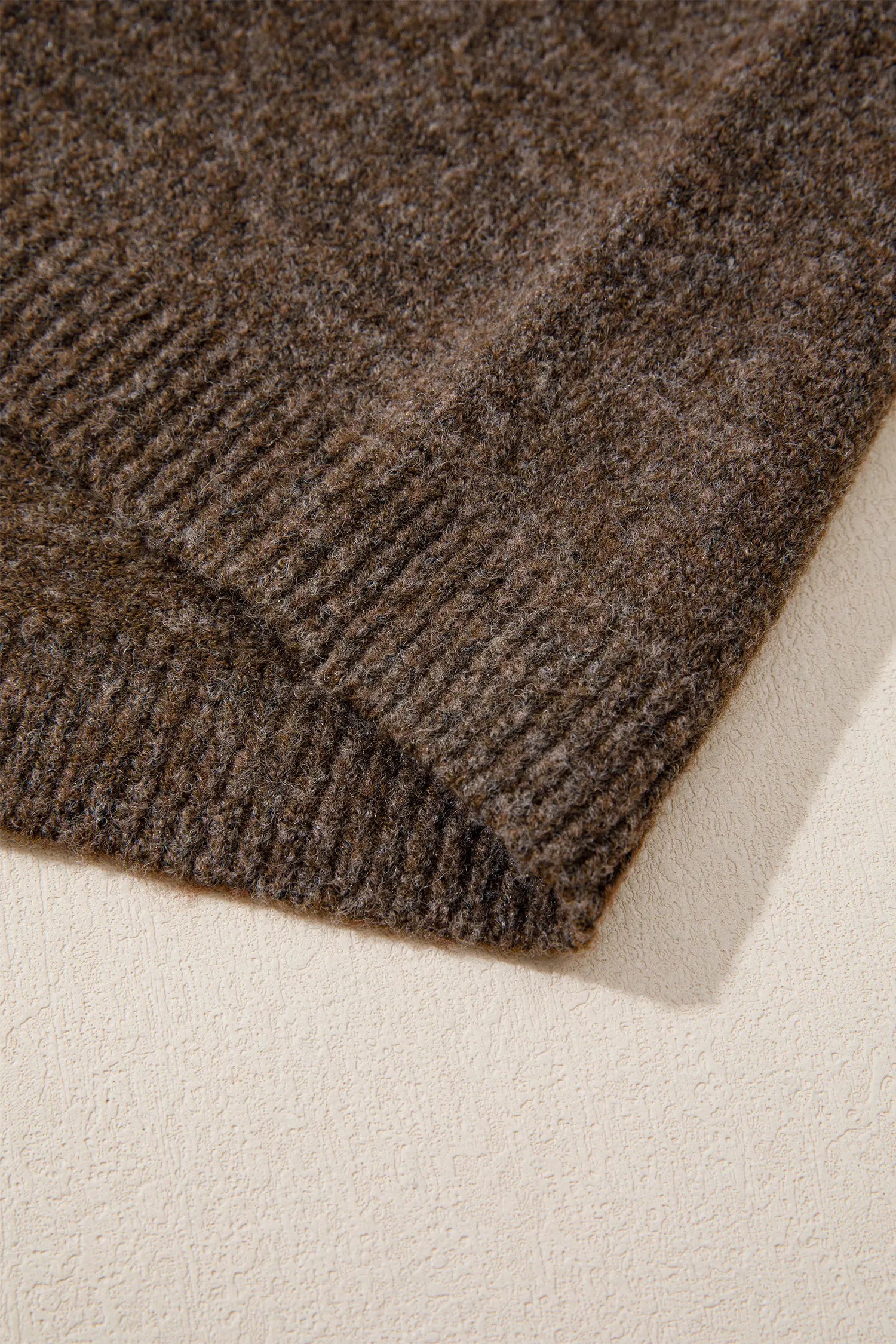 Close-up of a folded ribbed knit fabric in a heathered dark brown color, showing the texture and stitch detail against a light cream background.