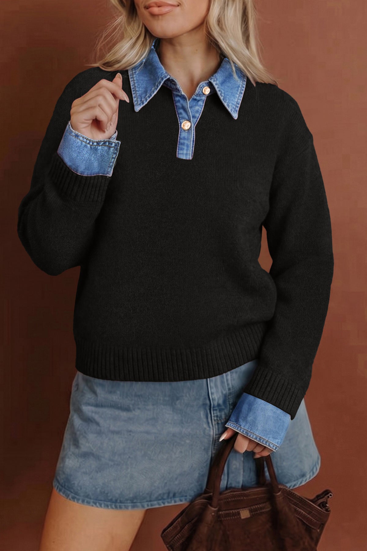 A woman wearing a black knit sweater with a light blue denim collar and matching denim button placket, paired with a light blue denim mini skirt and holding a brown handbag against a warm brown studio background.