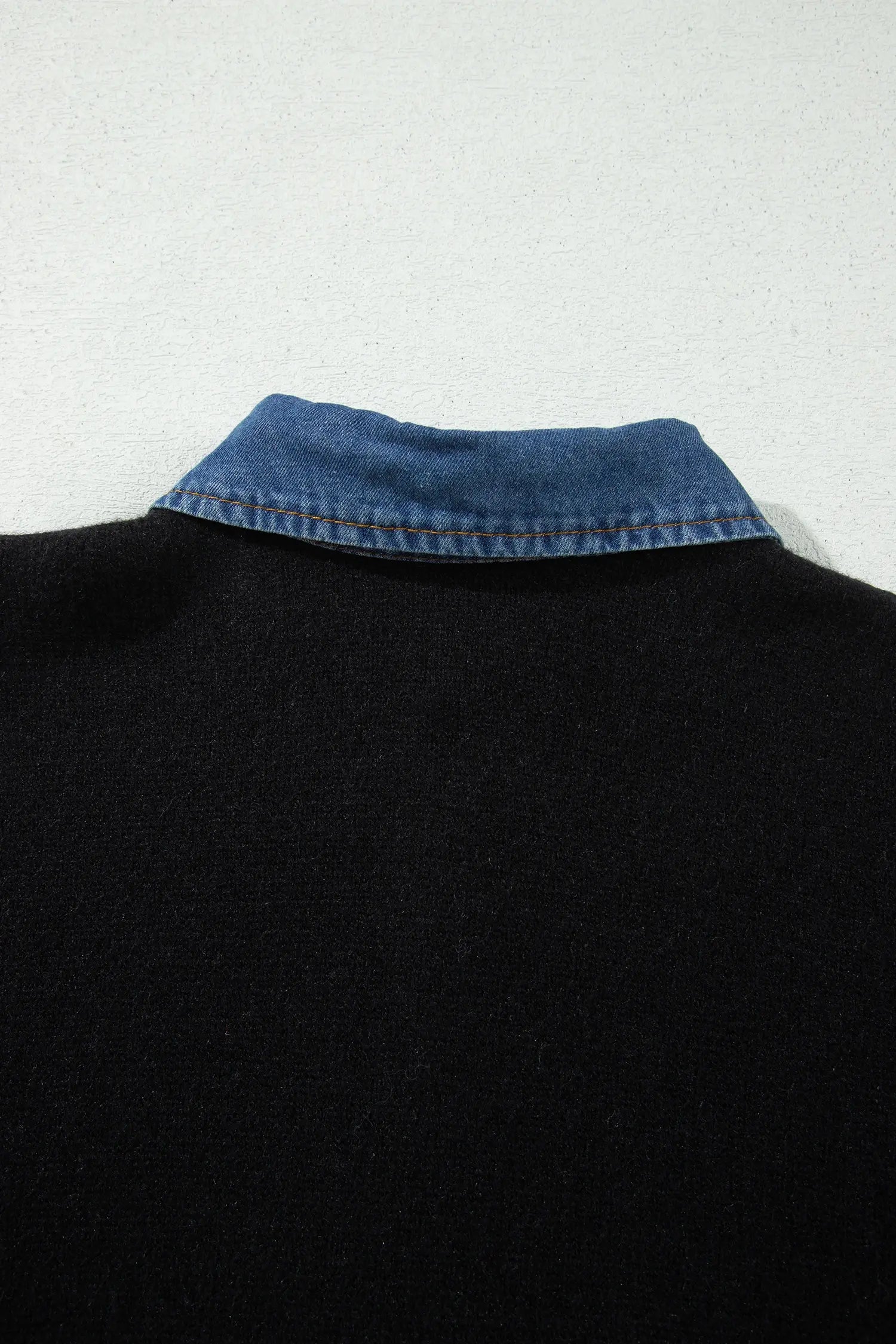 Close-up of the back of a black knit sweater with a contrasting blue denim collar, set against a light gray textured background.