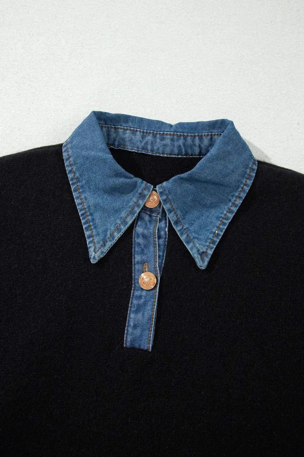 A close-up of a black knit polo-style top with a contrasting blue denim collar and matching denim placket, accented by two bronze buttons.