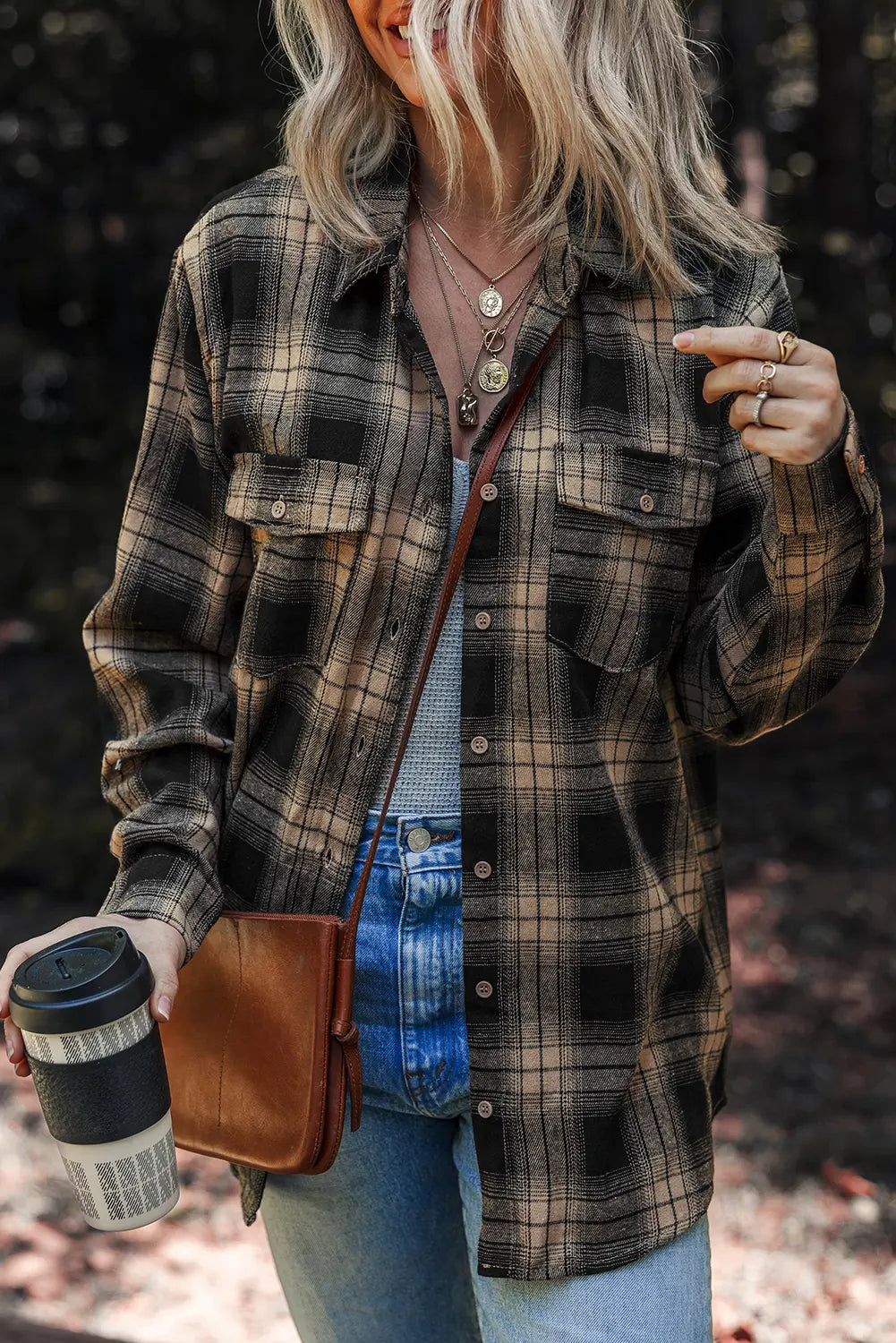 A woman wearing a brown and black plaid button-up flannel shirt over a light blue top and jeans, carrying a brown leather crossbody bag and holding a black travel coffee cup in an outdoor setting.