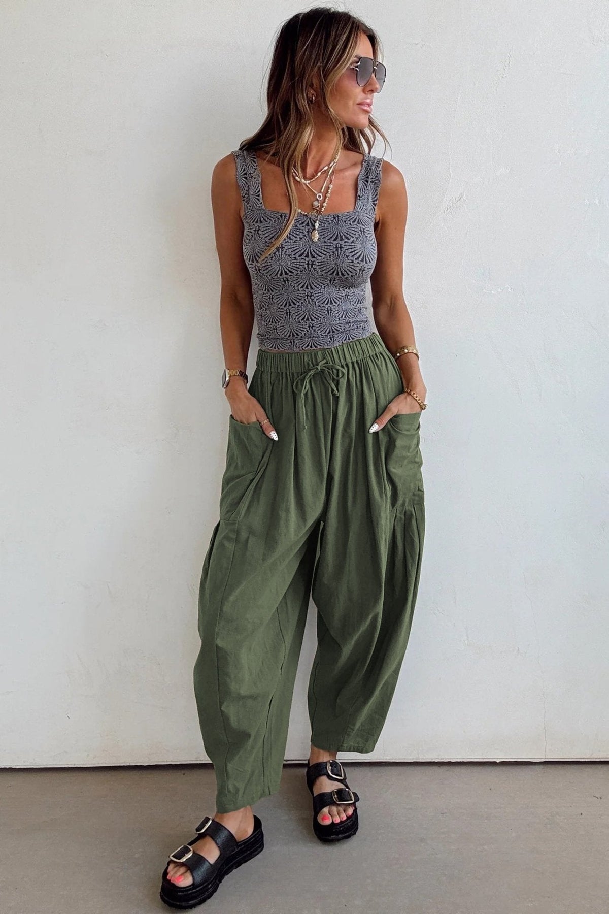 Woman wearing a gray tank top and green pants against a white wall.