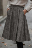A person wearing a gray and black herringbone patterned A-line skirt with side pockets, paired with a beige ribbed knit sweater and black leggings, standing outdoors on a paved street.