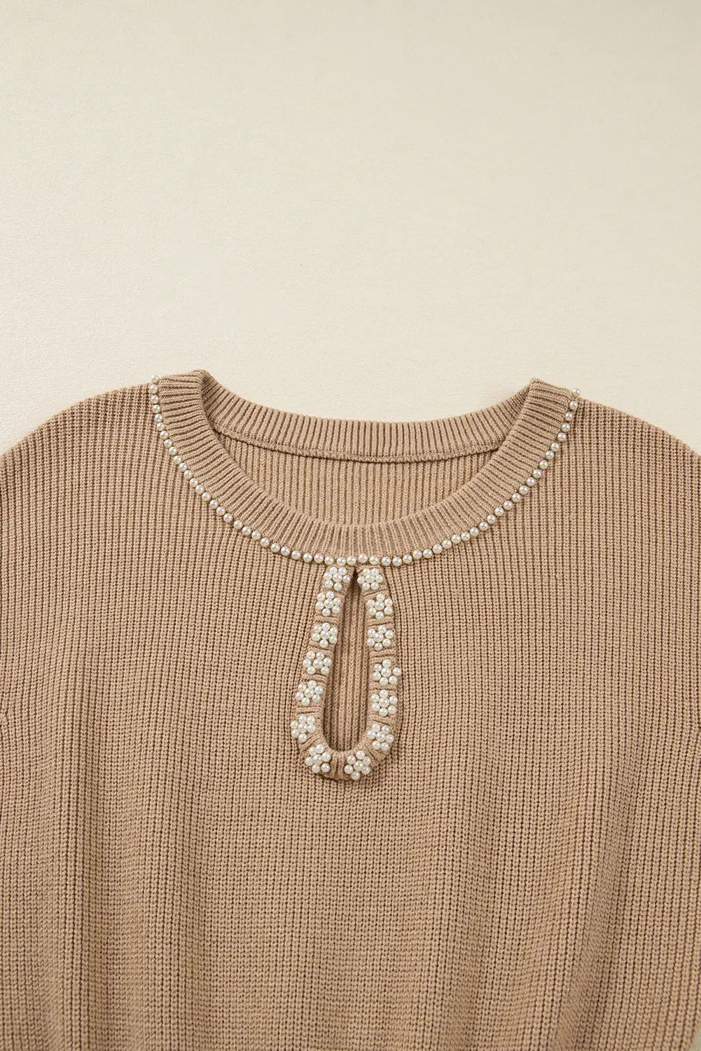 Close-up of a beige ribbed knit top with a decorative pearl and crystal keyhole neckline detail, set against a solid cream background.