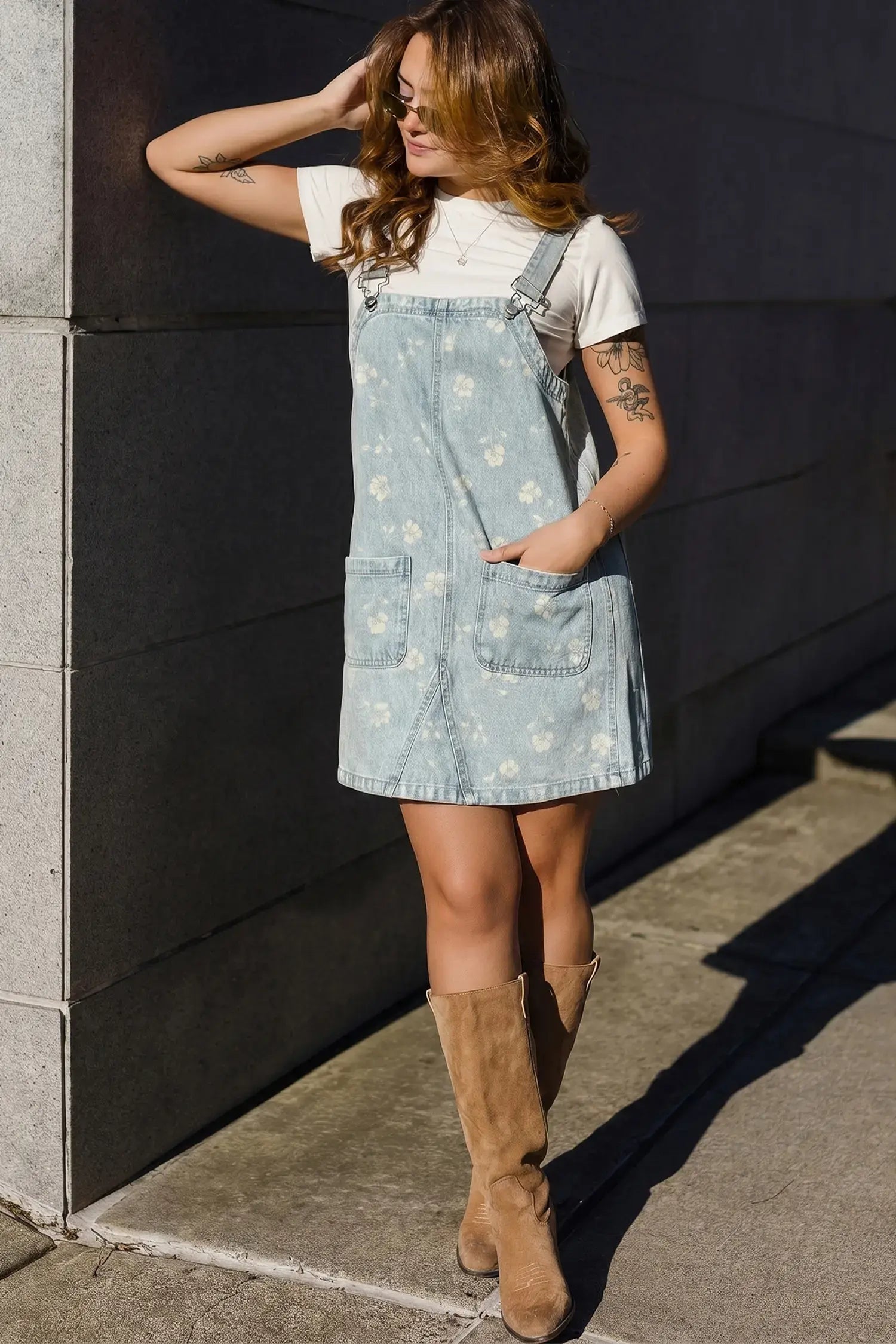 A woman wearing a light blue denim floral overalls dress over a white t-shirt and brown suede knee-high boots, posing outdoors against a dark gray concrete wall.