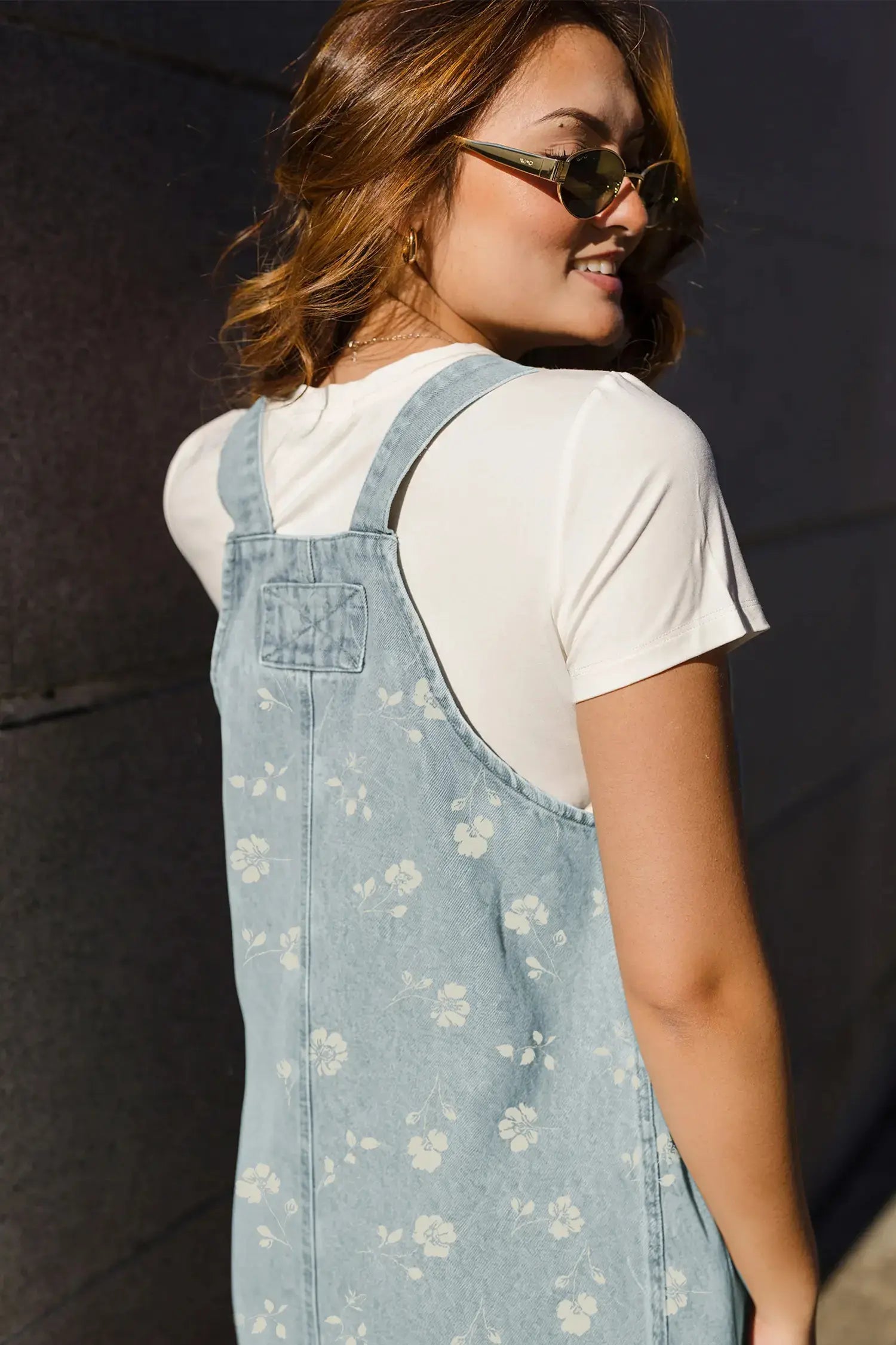A woman wearing a light blue denim overall dress with a white floral pattern over a white t-shirt, photographed from the back against a dark gray wall.