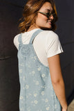 A woman wearing a light blue denim overall dress with a white floral pattern over a white t-shirt, photographed from the back against a dark gray wall.