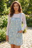 A woman wearing a light blue denim overall dress with a white floral pattern over a white long-sleeve shirt, standing outdoors on a stone path with green foliage in the background.