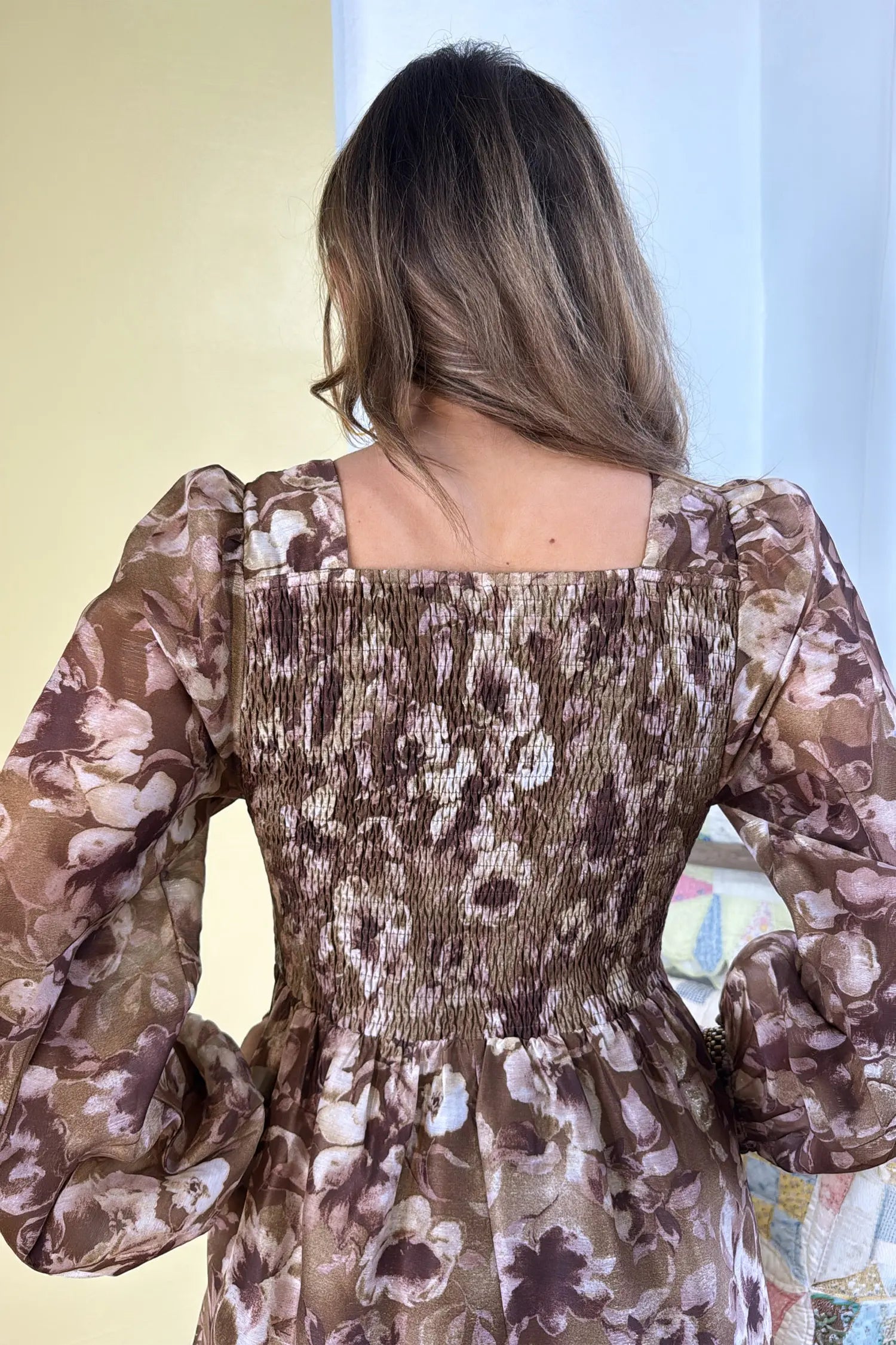 A woman viewed from behind wearing a brown floral smocked dress with long puff sleeves and a square neckline.