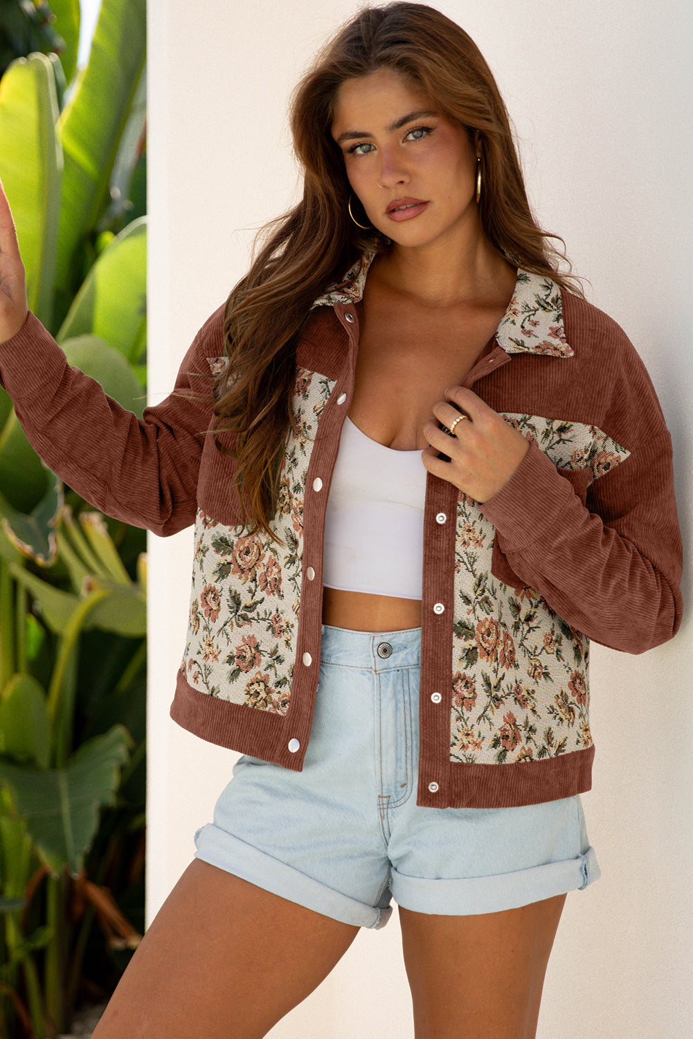 Woman wearing a floral and brown jacket with denim shorts against a white wall.