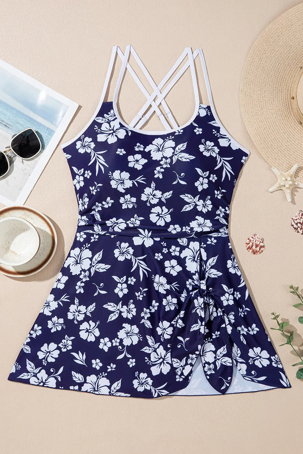 Navy blue floral dress with white straps on a beige surface with a hat and sunglasses.