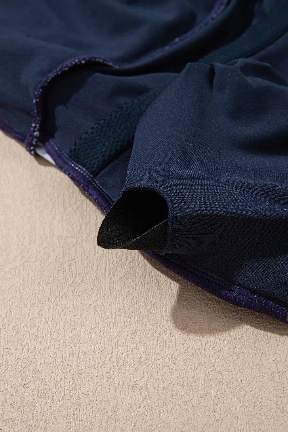 Close-up of a dark navy blue garment, likely a swimsuit or athletic top, showing the fabric texture, mesh panel, and dark piping along the edge, laid on a textured beige surface.