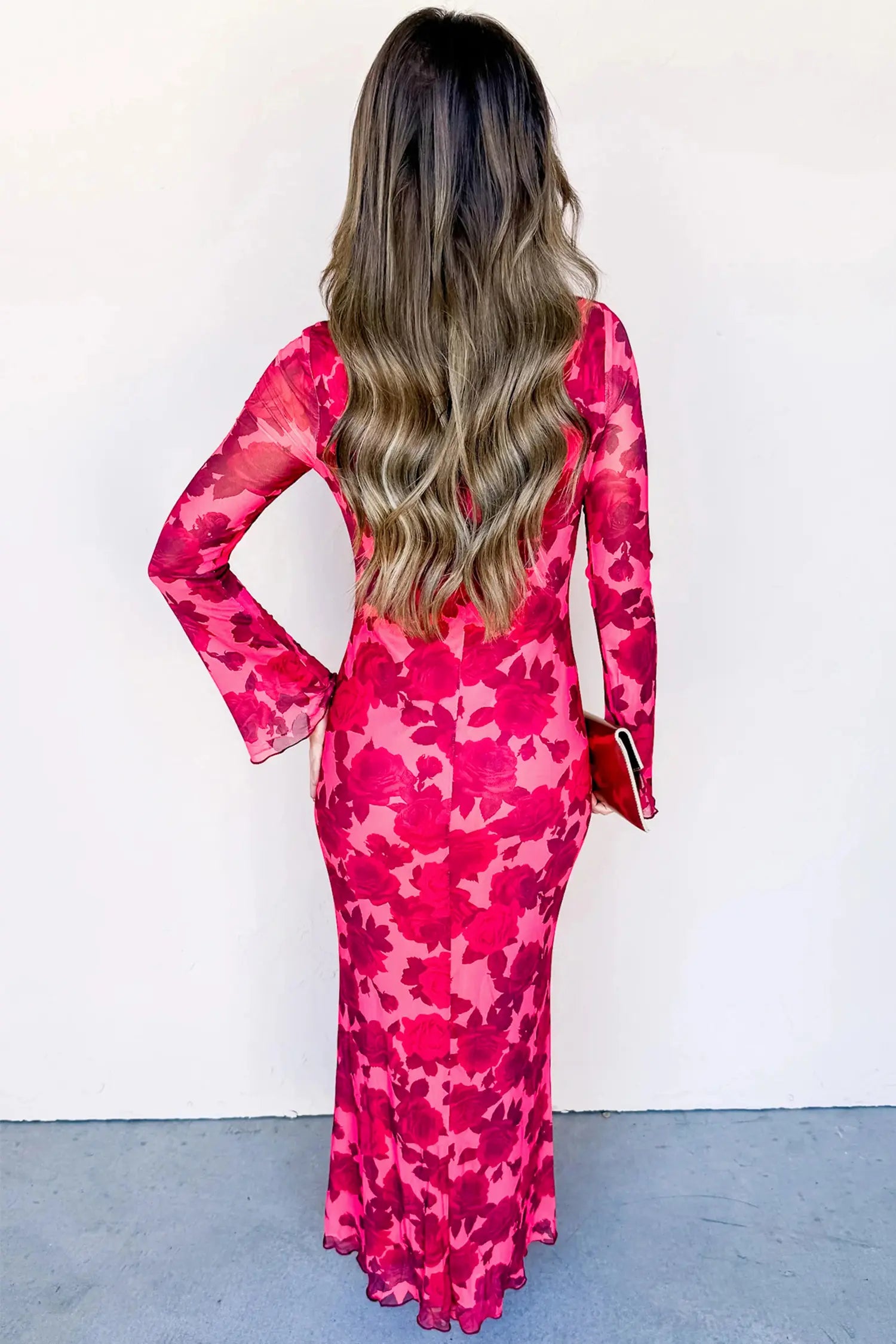 A woman viewed from behind wearing a long-sleeve, form-fitting maxi dress with a vibrant pink and red floral pattern. She is holding a small red clutch against a plain white wall and gray floor.