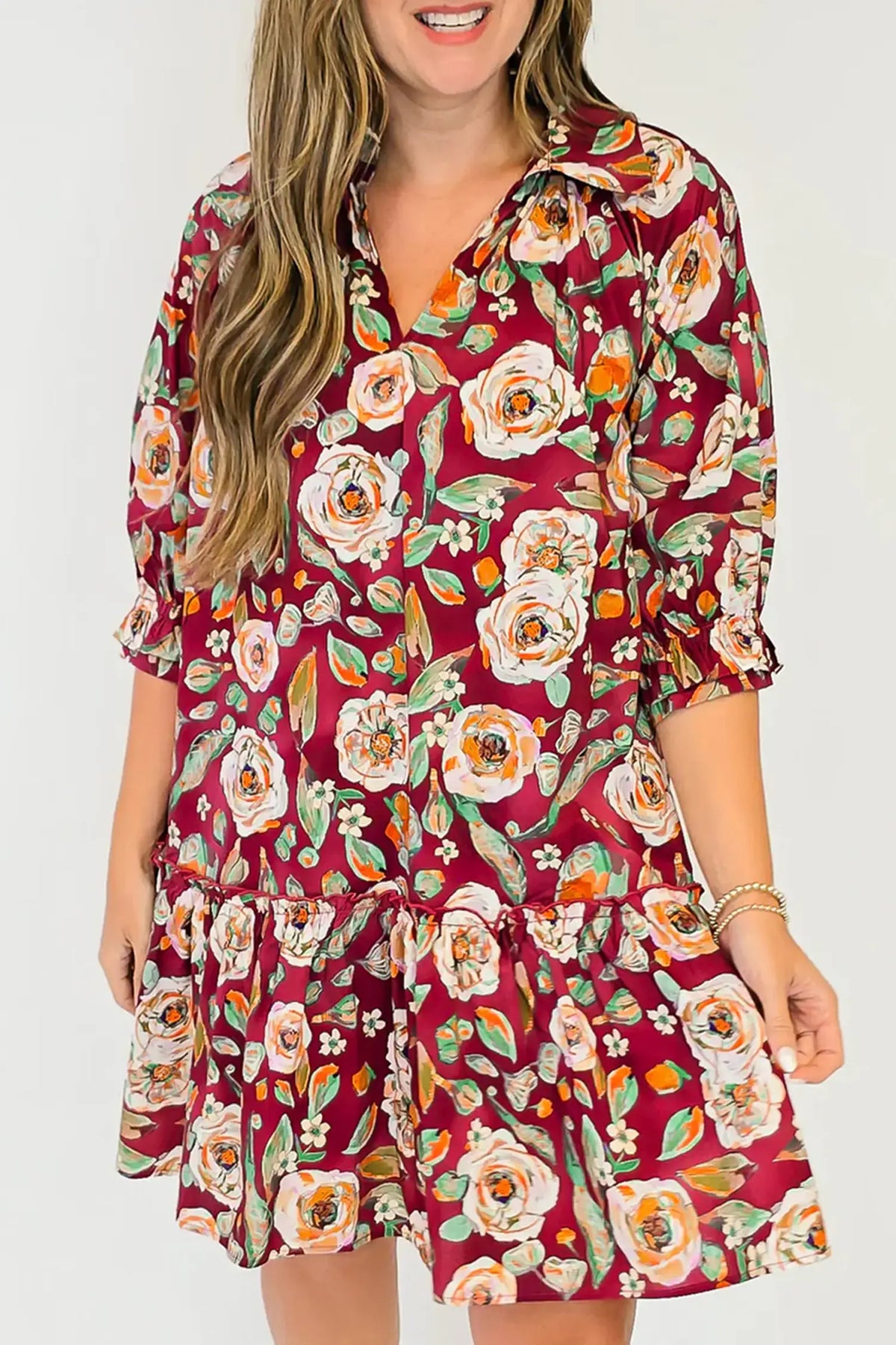 A woman wearing a short-sleeve collared dress with a red base and a large floral print in white, orange, and green, shown against a plain light gray background.