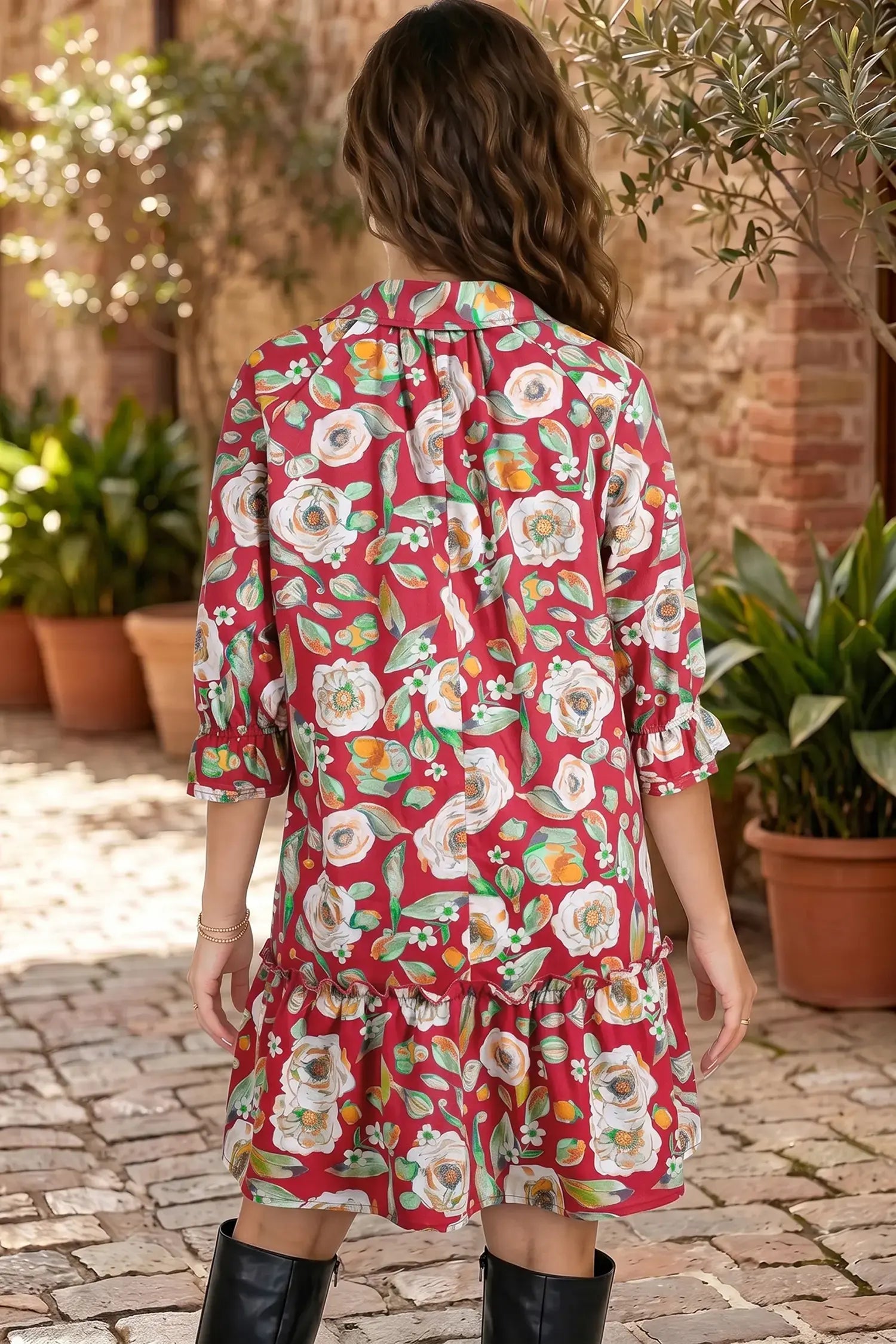 A woman viewed from behind wearing a red tiered shirt dress with a colorful floral print, standing on a cobblestone patio with potted plants and a brick wall in the background.