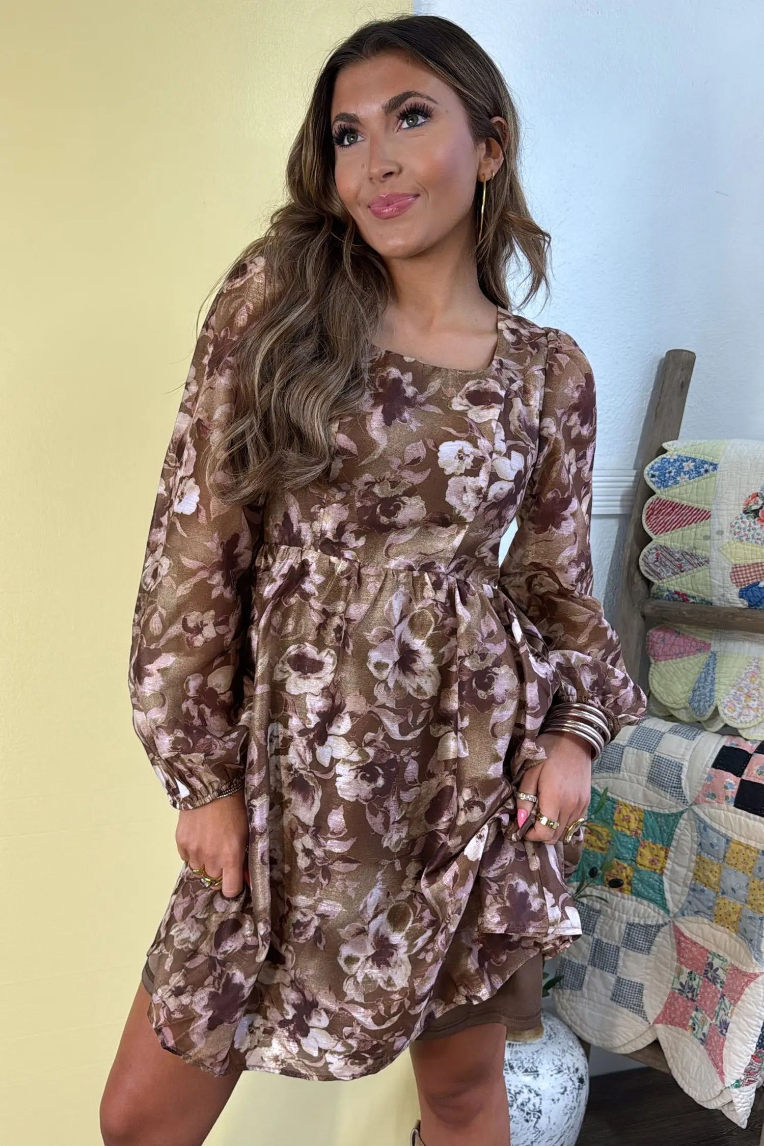 A woman wearing a brown and beige floral long-sleeve mini dress, standing in front of a pale yellow wall with a quilted chair visible in the background.