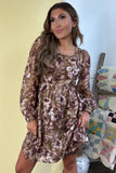 A woman wearing a brown and beige floral long-sleeve mini dress, standing in front of a pale yellow wall with a quilted chair visible in the background.