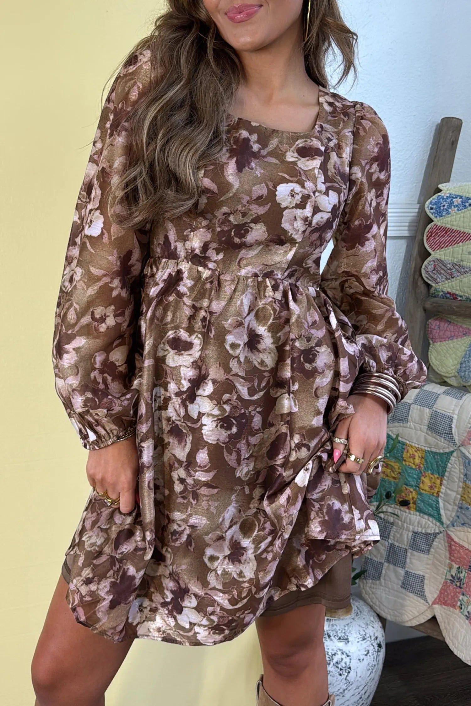 A woman wearing a brown floral long-sleeve mini dress with a square neckline and gathered waist, standing indoors next to a quilted chair.