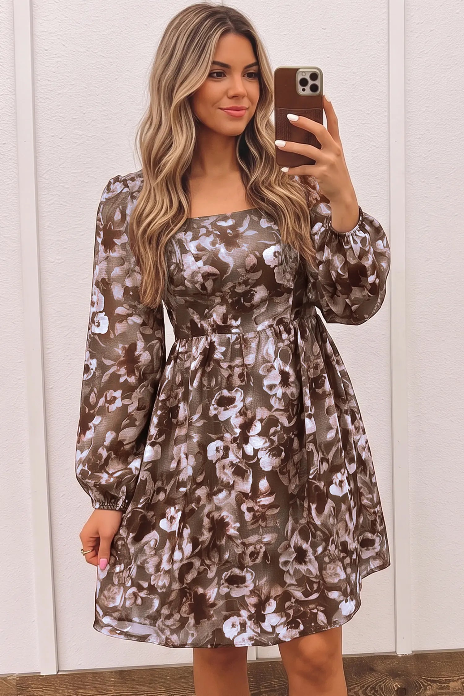 A woman takes a mirror selfie wearing a long-sleeve, knee-length dress with a brown, beige, and white floral pattern. The dress features a square neckline, a fitted waist, and a flared skirt, set against a white paneled wall.