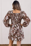 A woman viewed from the back wearing a brown floral puff-sleeve dress with a smocked bodice and square neckline, standing against a light paneled wall.
