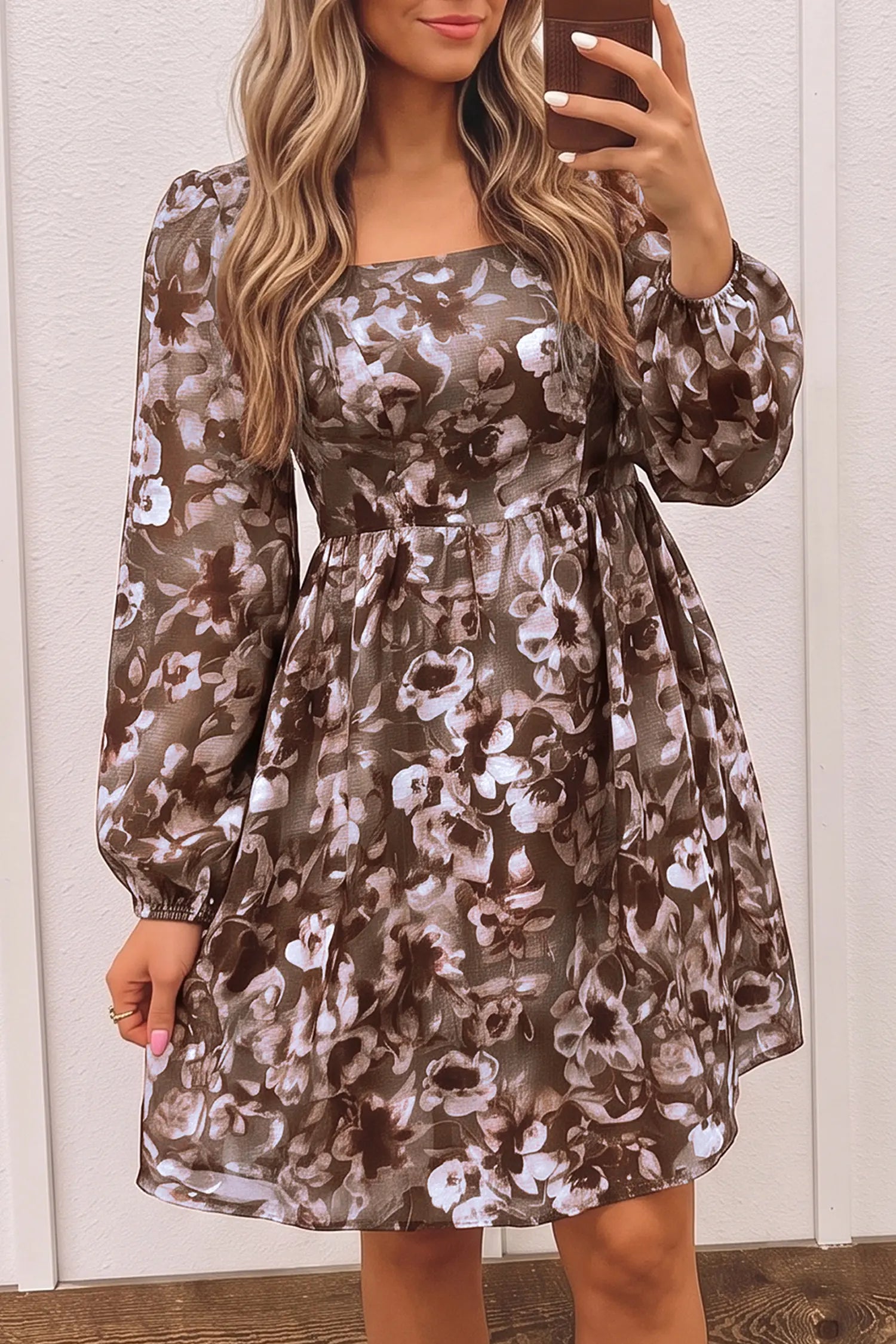 A woman takes a mirror selfie showing a long-sleeve, knee-length dress with a brown, beige, and white floral pattern. The dress features a square neckline, long balloon sleeves, and a gathered waist.