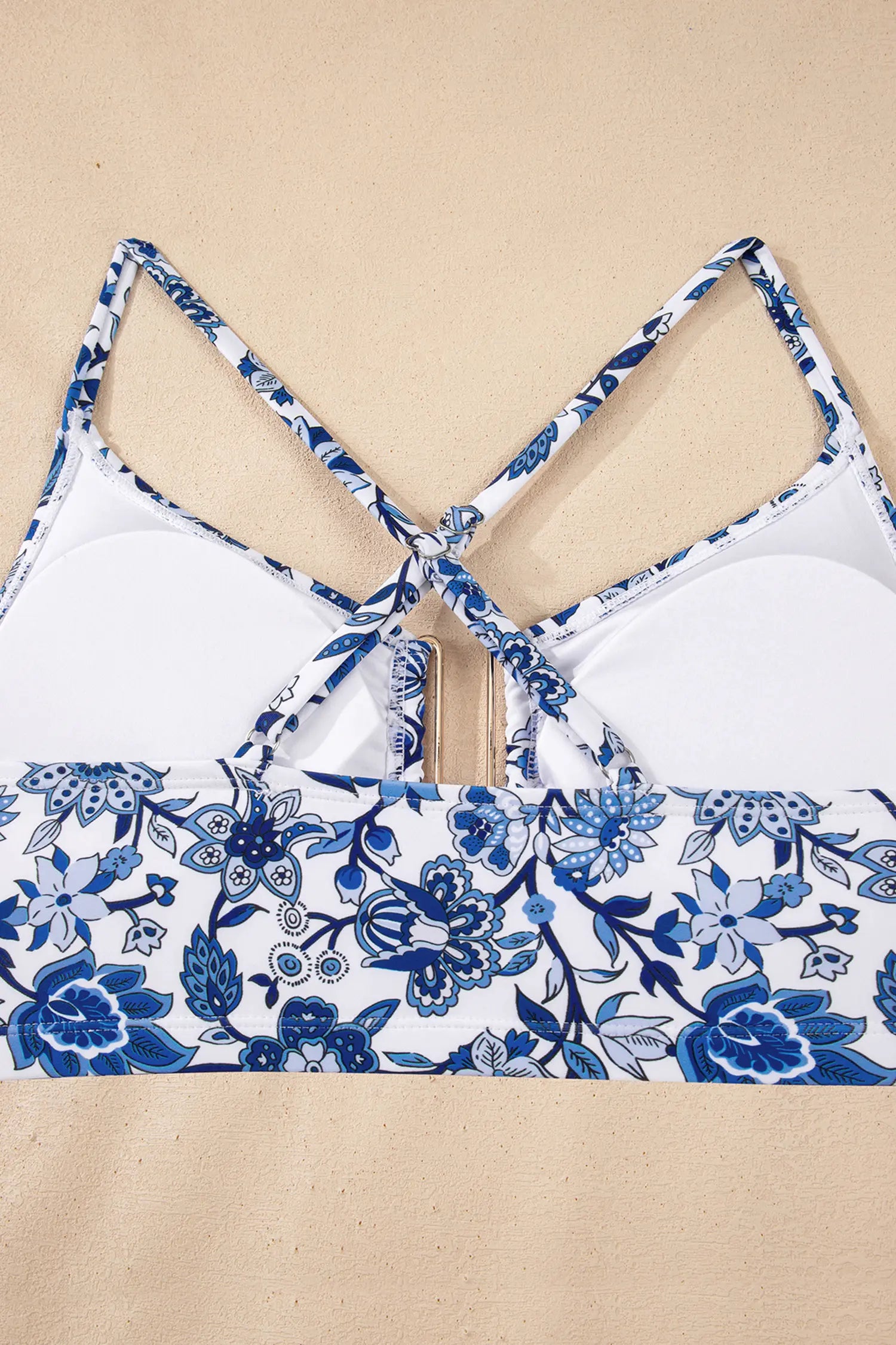A close-up of a bikini top or bralette laid flat on a beige surface, showing white cups with blue floral print straps and a matching blue floral band across the lower front.