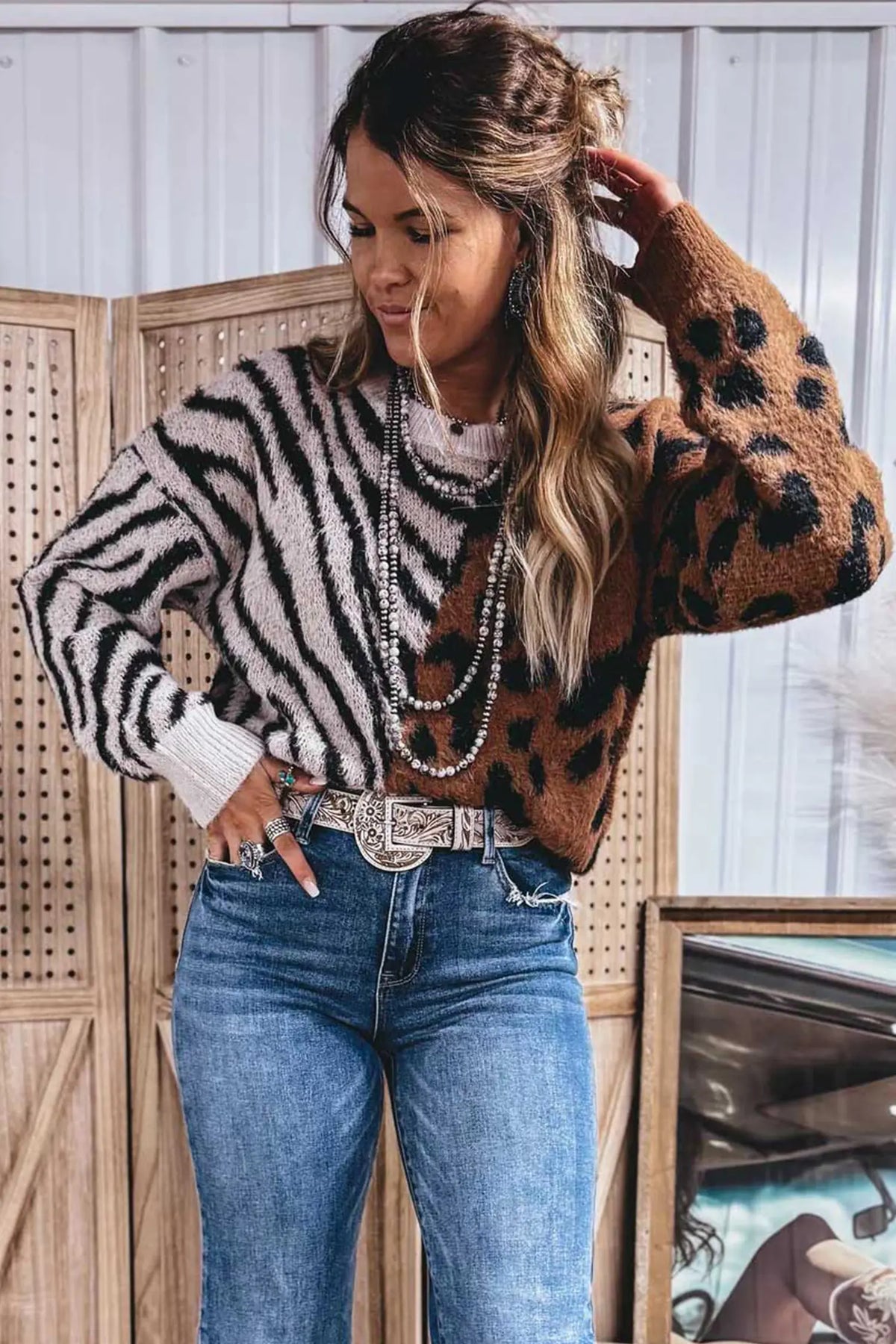 A woman models a cropped knit sweater featuring a split design: one half is black and white zebra print, and the other half is brown with black leopard spots. She is styled with blue jeans, a silver belt, and layered pearl necklaces, standing in front of a wooden screen.